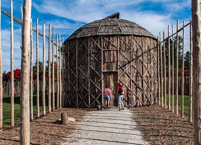 Round thatched wooden hut with crisscross lattice, approached by a path lined with tall wooden posts; people stand at the entrance.