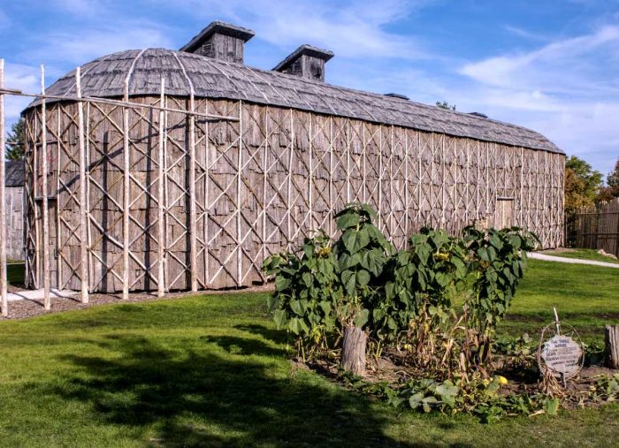 Exterior view of a long, wooden lattice-structured building with a rounded shingled roof on a well-kept lawn.