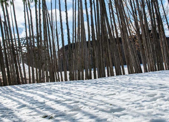 Snow-covered ground in a yard with tall, closely spaced wooden fence posts and a dark cabin behind them.