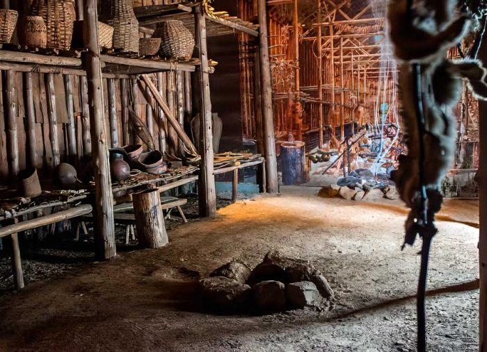 Interior of a rustic workshop with wooden shelves, baskets, pots, and a central fire pit.