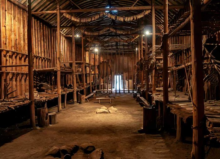 Dimly lit wooden interior of a rustic workshop or storage hall with long benches and shelves along both sides.