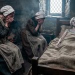 Two women in old-fashioned dress sit by a stone bed, covering their faces in prayer beside a bedridden woman in a rough blanket. A candle and small window illuminate the dim room.